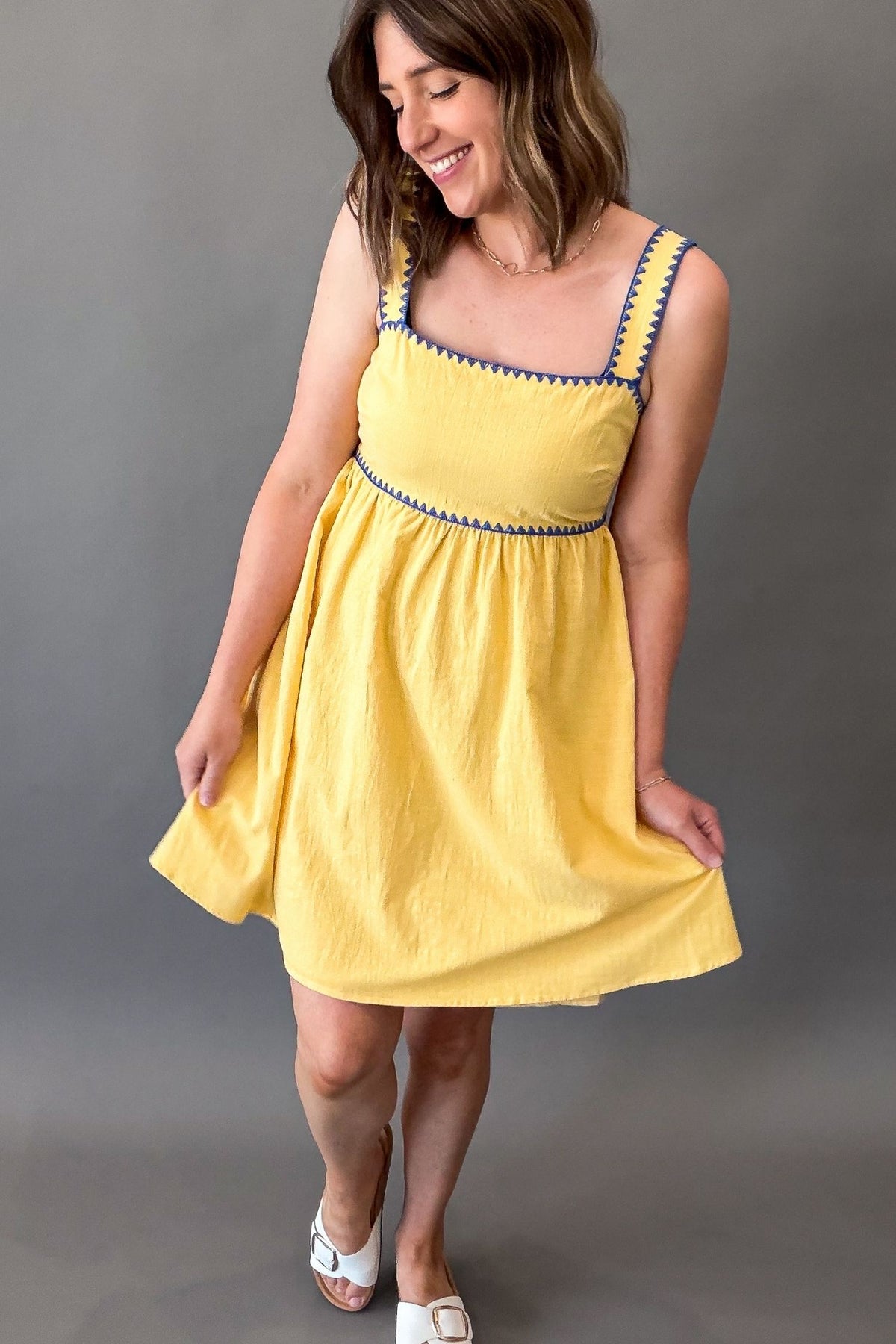 Woman wearing a sunny yellow midi dress featuring a square neckline, blue contrast trim, adjustable shoulder straps, tie-back detail, side pockets, and a flowy lined skirt.