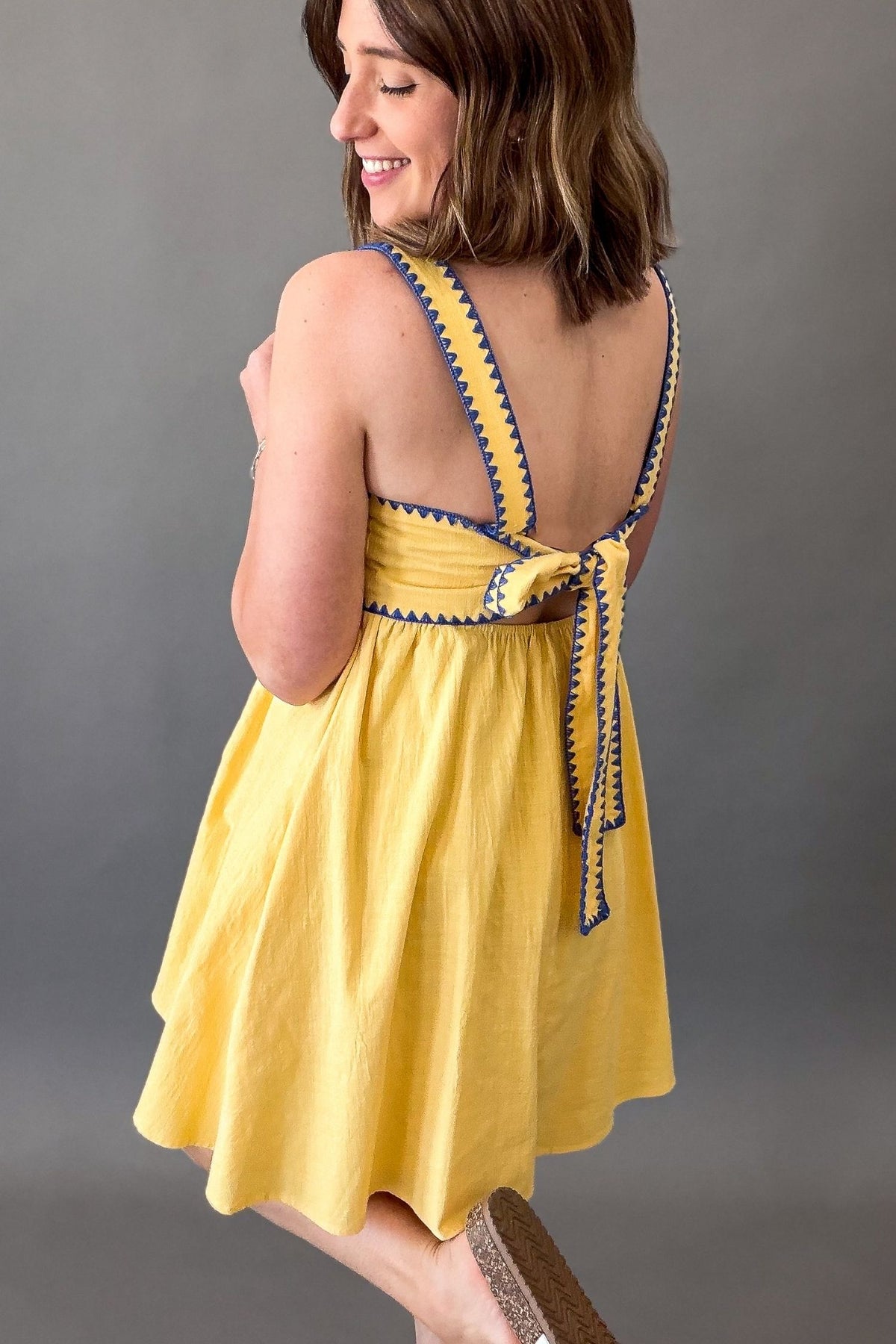 Woman wearing a sunny yellow midi dress featuring a square neckline, blue contrast trim, adjustable shoulder straps, tie-back detail, side pockets, and a flowy lined skirt.
