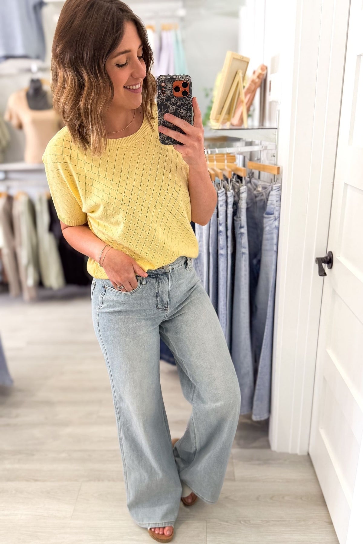 Woman taking a mirror selfie wearing a yellow top and light blue jeans in a clothing store.