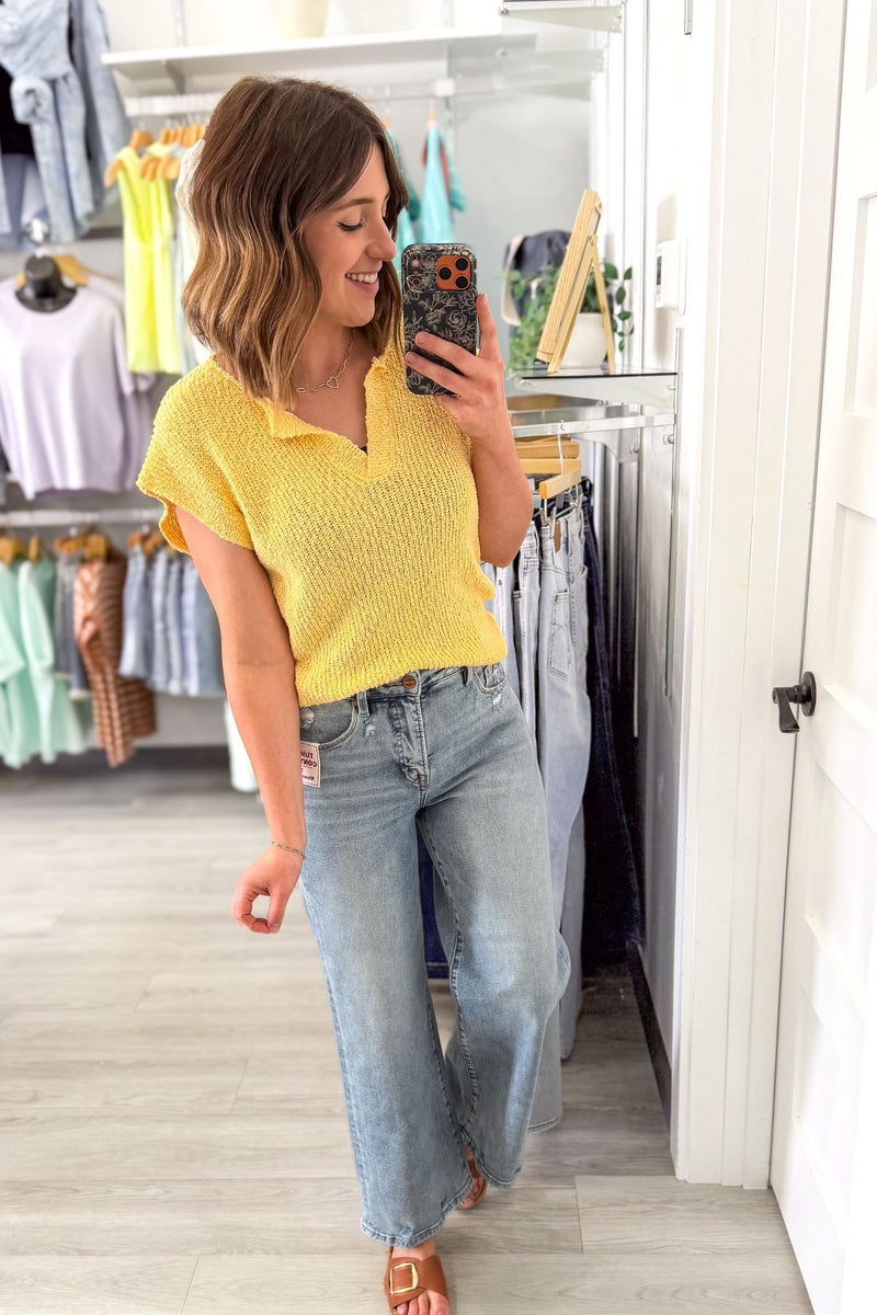 Woman taking a mirror selfie wearing a yellow top and blue jeans in a clothing store.